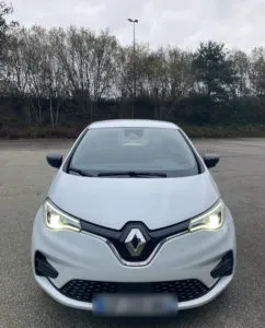 Voiture électrique Renault Zoé blanche, vue de face avec phares allumés, stationnée sur un parking